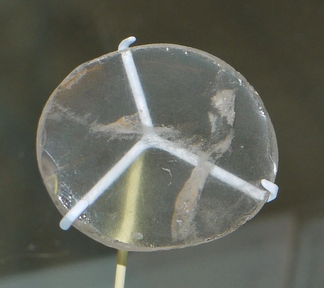 A clear, round ancient Roman glass disk with a white "X" pattern, held upright by two support clips on its edge, displayed against a dark background.