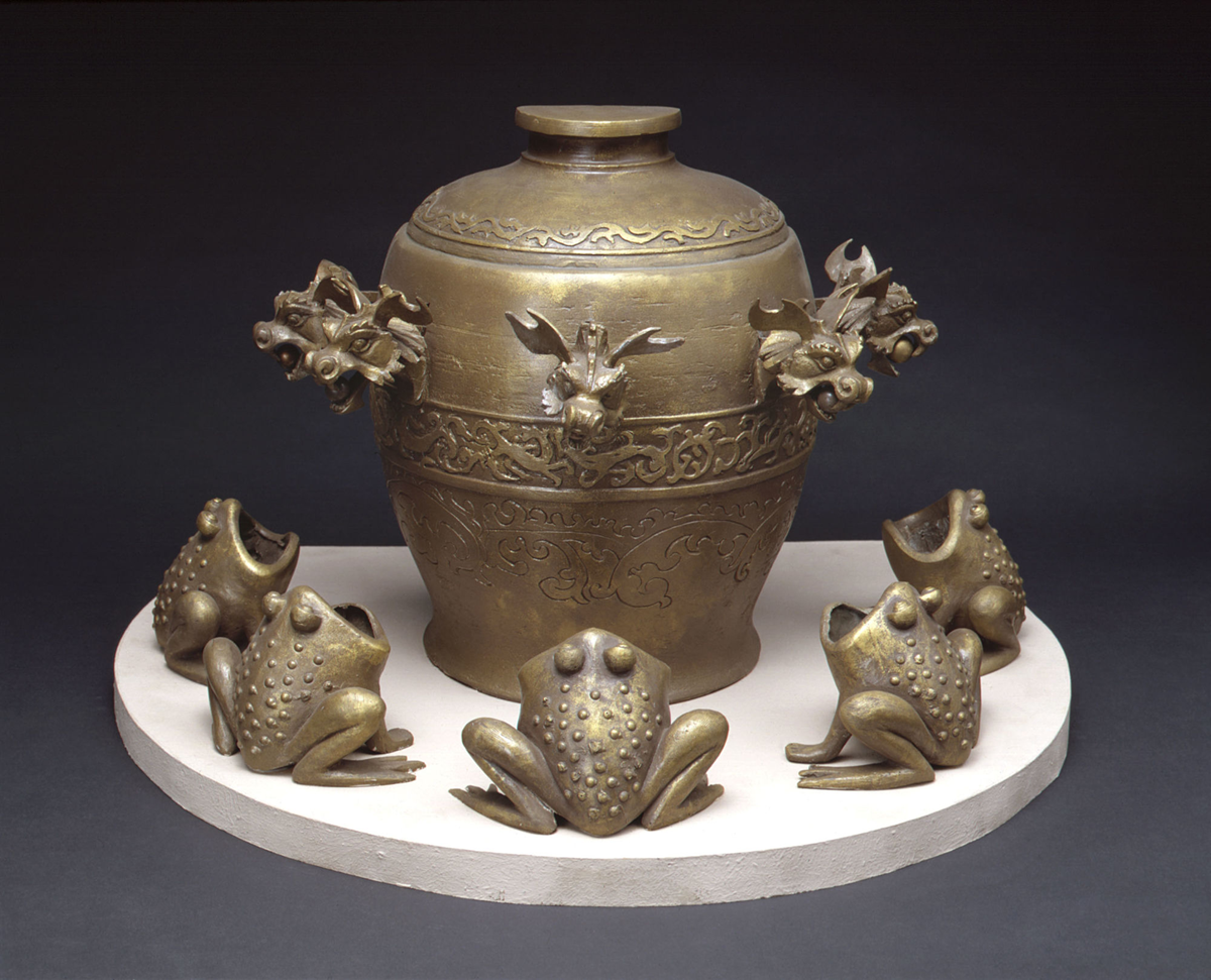 A bronze jar with ornate dragon heads as handles, surrounded by six frog-shaped figurines, all placed on a round, white platform against a dark background.