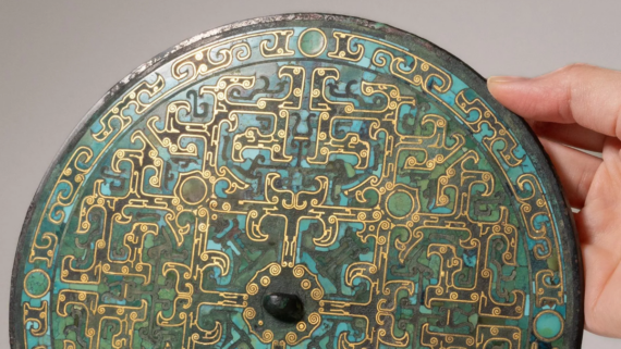 A hand holds a round, ornate bronze mirror with intricate gold and turquoise patterns, geometric designs, and a central knob on its decorated back.