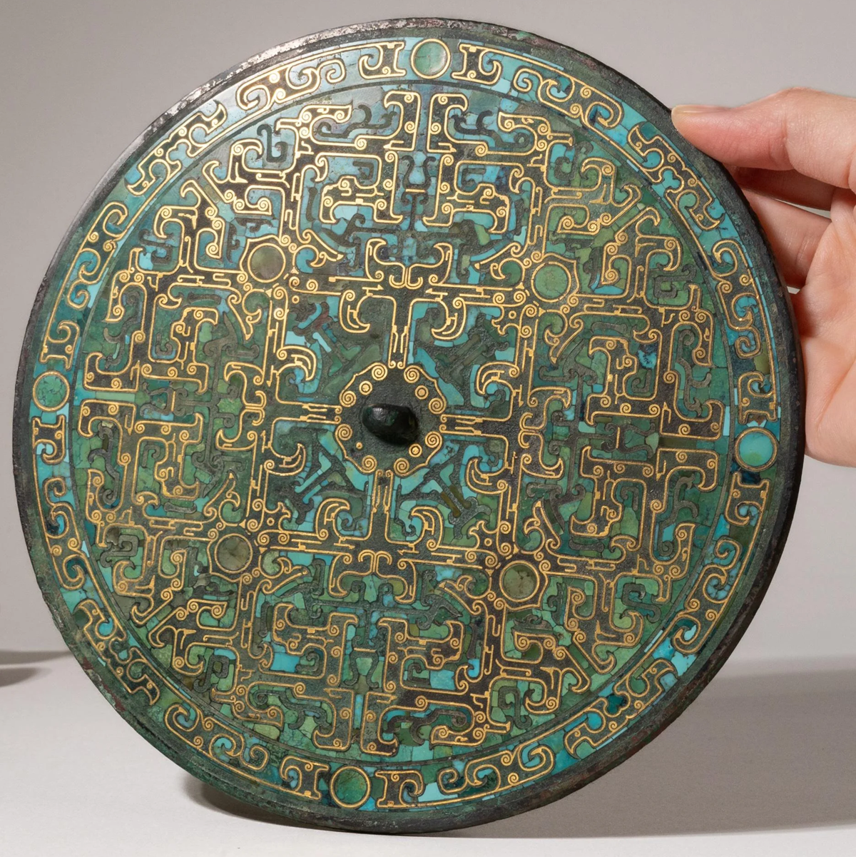 A hand holds a round, ornate bronze mirror with intricate gold and turquoise patterns, geometric designs, and a central knob on its decorated back.