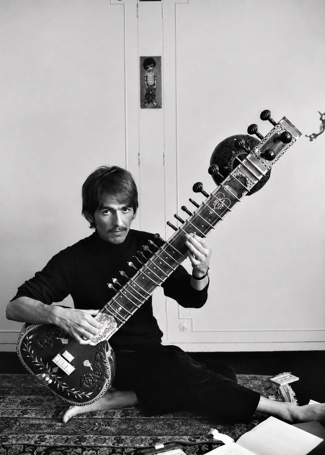 A man sits cross-legged on the floor, playing a sitar. He has short hair and wears dark clothing. The room is simply decorated, with a patterned rug and a small framed picture on the wall behind him.