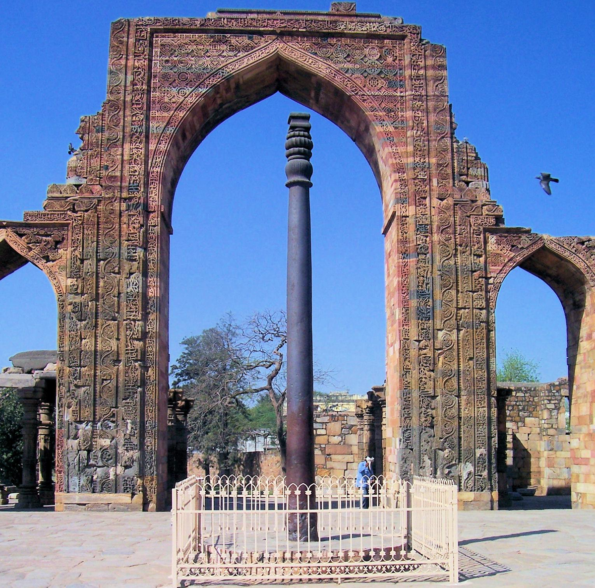 The Iron Pillar of Delhi stands protected by a metal fence in the foreground, framed by the ornate, carved stone archway of the Quwwat-ul-Islam Mosque at the Qutub Minar complex under a bright blue sky. A bird is in flight to the right.