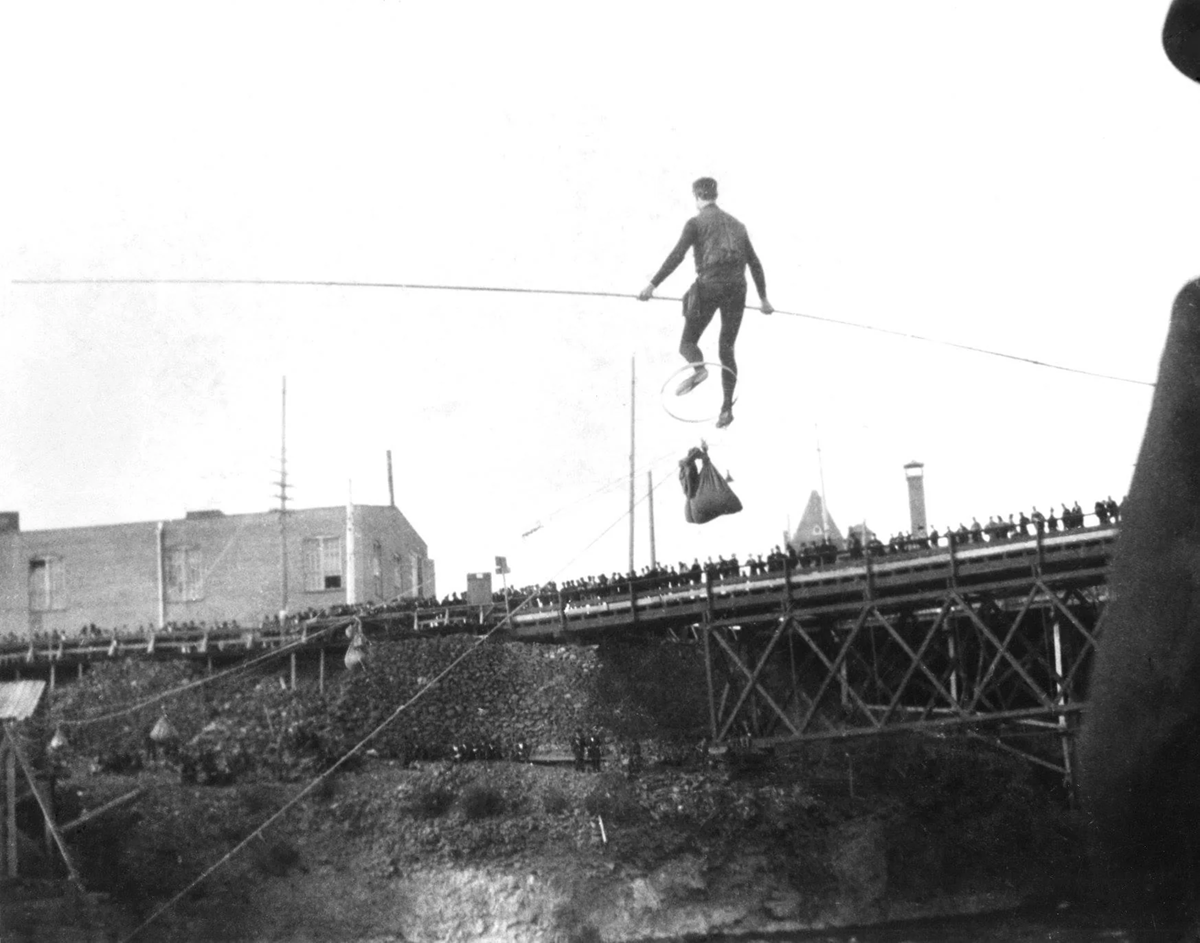 A person walks on a tightrope over a river, carrying items and balancing with a wheel, while a large crowd watches from a nearby bridge and riverbank.