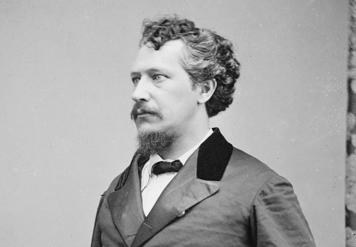 Black-and-white portrait of a man with curly hair, mustache, and goatee, wearing a double-breasted coat with black collar and a white shirt, looking slightly to the side against a plain background. Figure