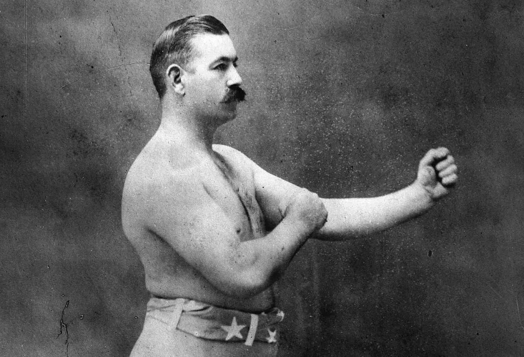 A vintage black-and-white photo of a mustached, shirtless man in boxing shorts, standing in profile and raising his fists in a classic boxing stance against a plain background.