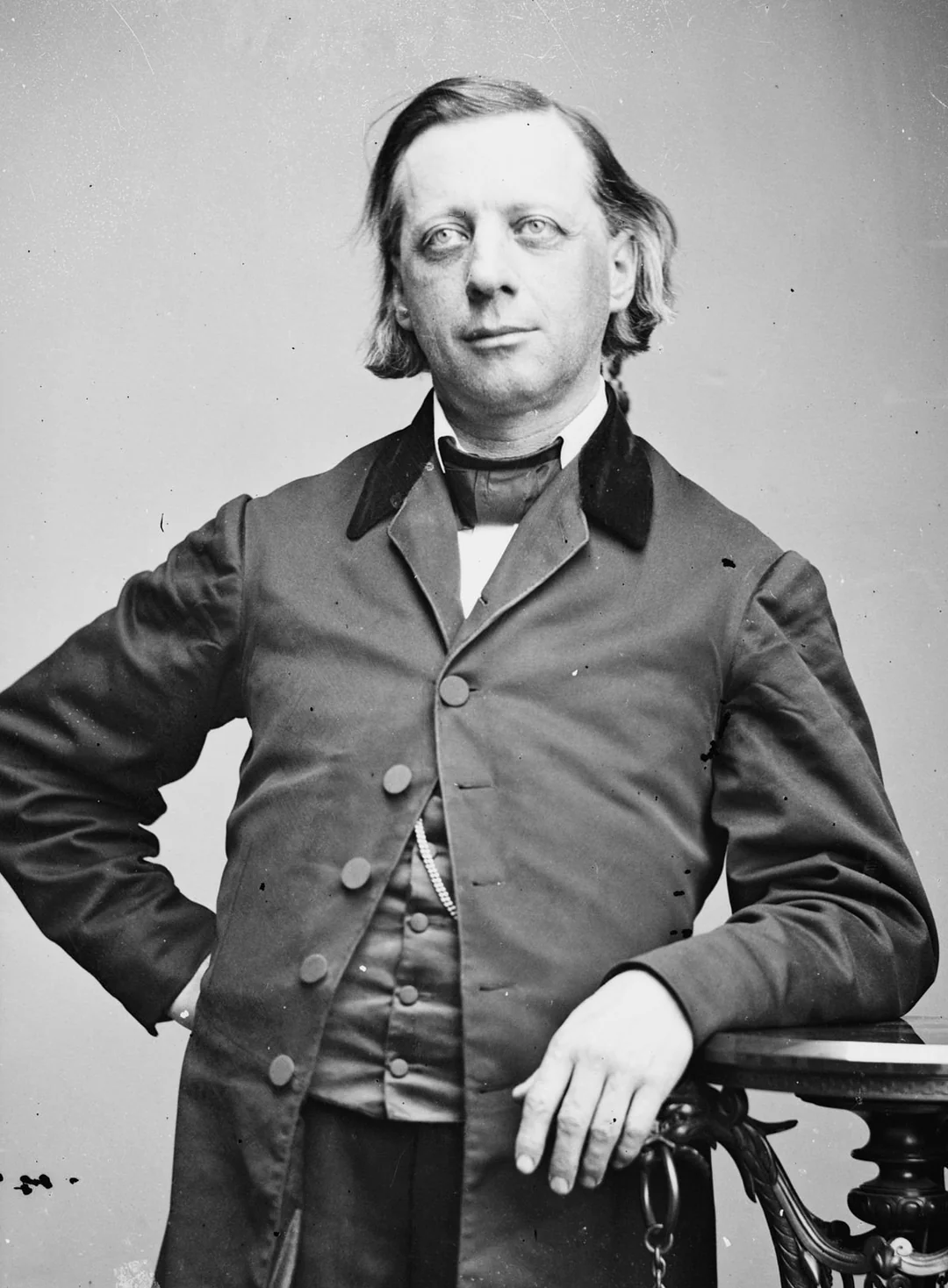 A man with light eyes and medium-length hair poses in a formal suit with one hand on his hip and the other resting on a wooden table. The photograph is black and white and appears to be from the 19th century.