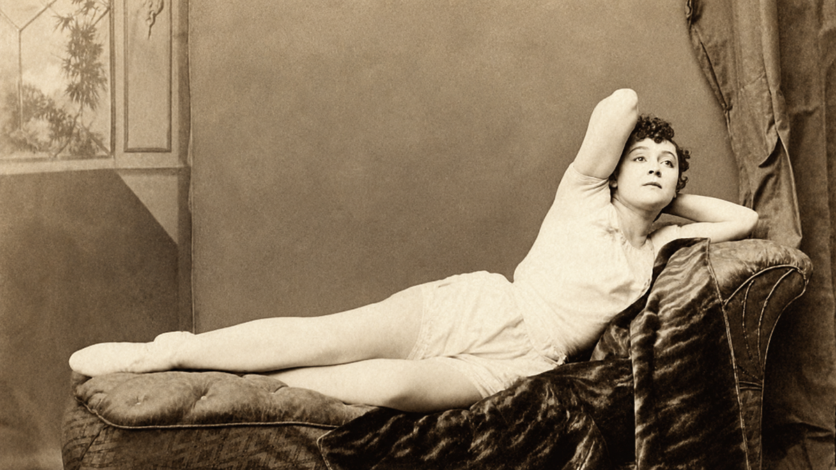 Sepia photo of a woman in a light outfit reclining on a chaise lounge, resting her head on one arm and stretching out her legs, gazing upward. Velvet fabric drapes beside her and a window is visible in the background.