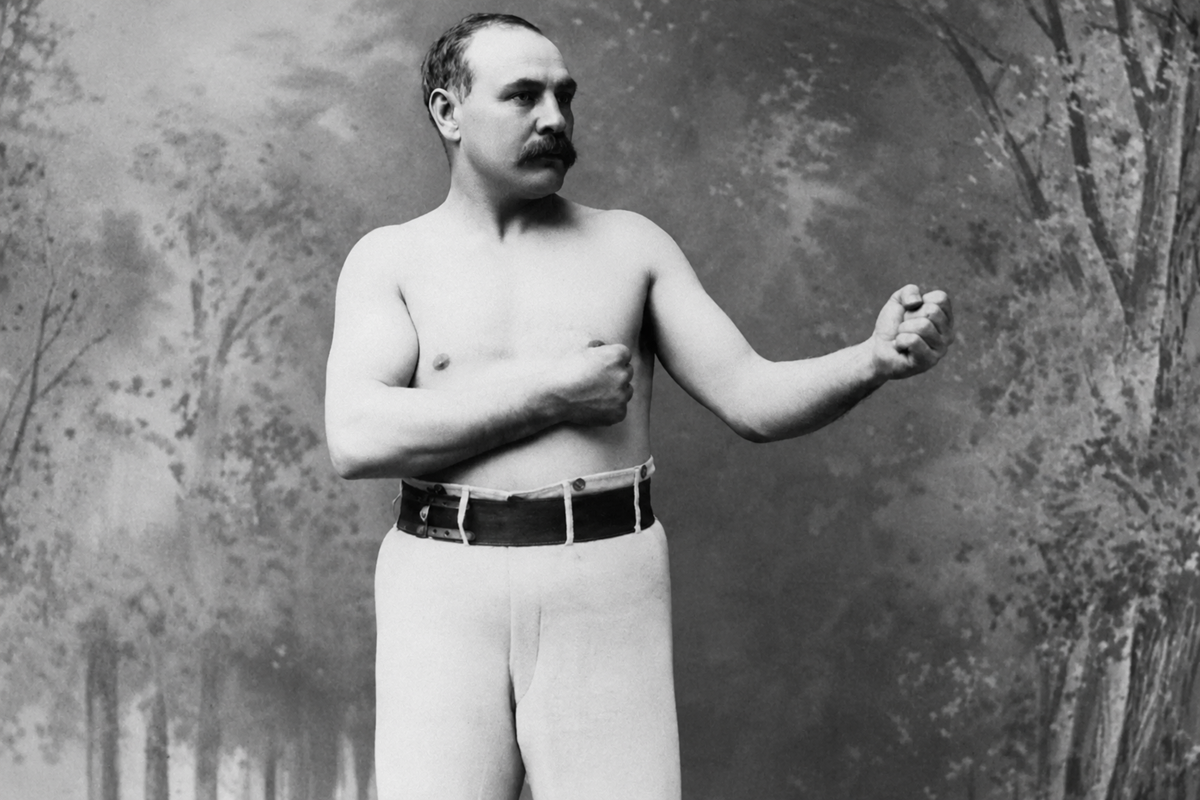 Black-and-white photo of a mustached man posing shirtless with fists raised, wearing light pants and a dark belt, against a painted backdrop of trees.
