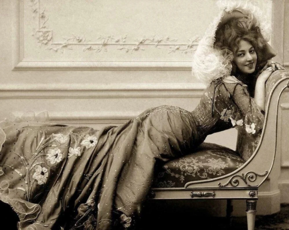A woman in an elegant, embroidered gown and large feathered hat reclines on a vintage chaise lounge, smiling at the camera in a vintage, sepia-toned setting.