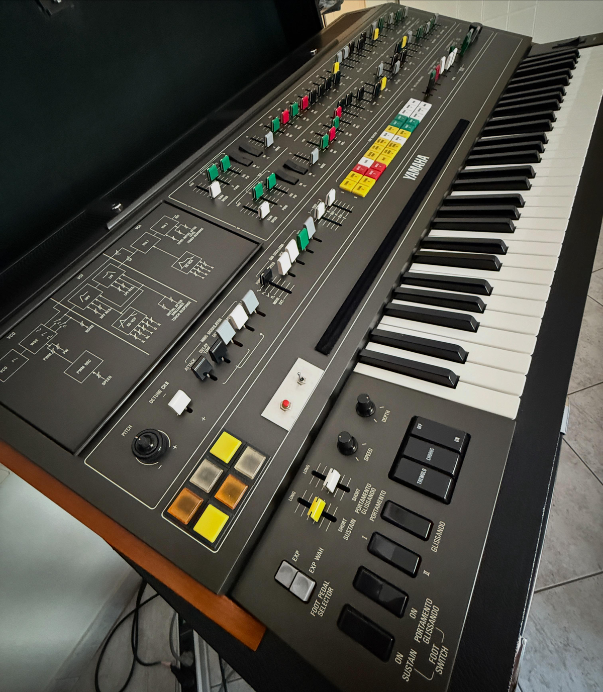 A close-up of a vintage Yamaha CS-80 synthesizer keyboard, showing its various colored buttons, sliders, switches, and keys on a dark panel with circuit diagrams.