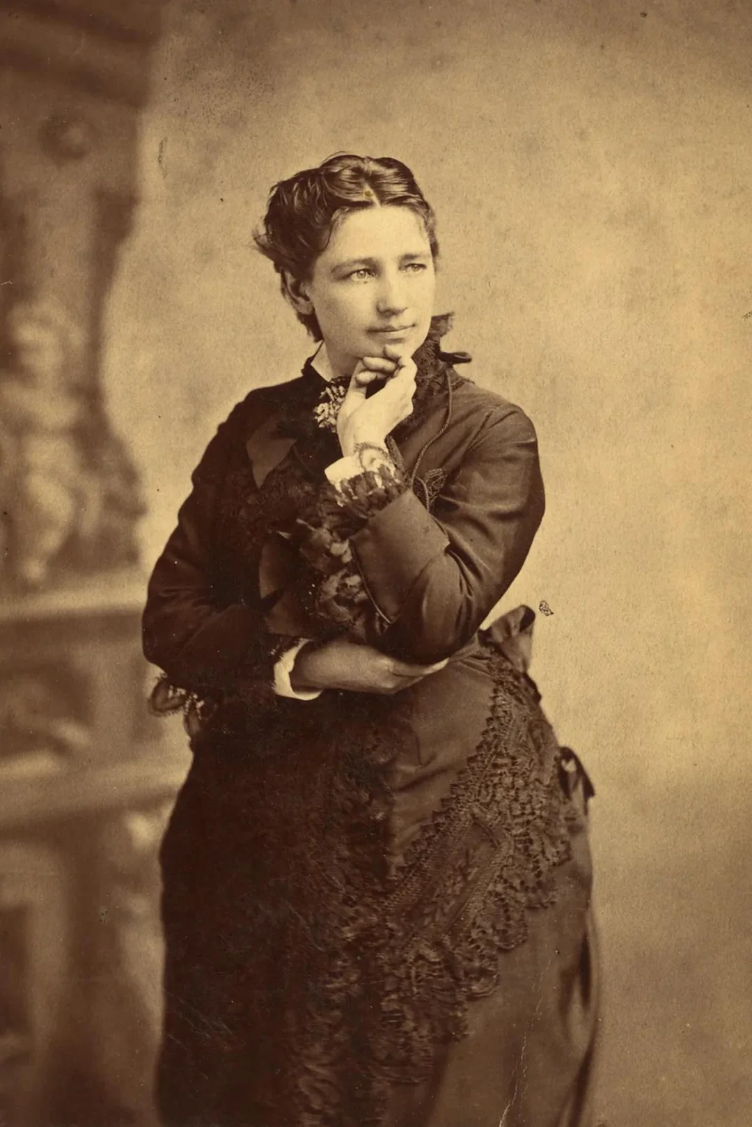 A woman in Victorian-era clothing poses with one hand on her chin, looking thoughtfully to the side. The sepia-toned photo has a soft, vintage background.