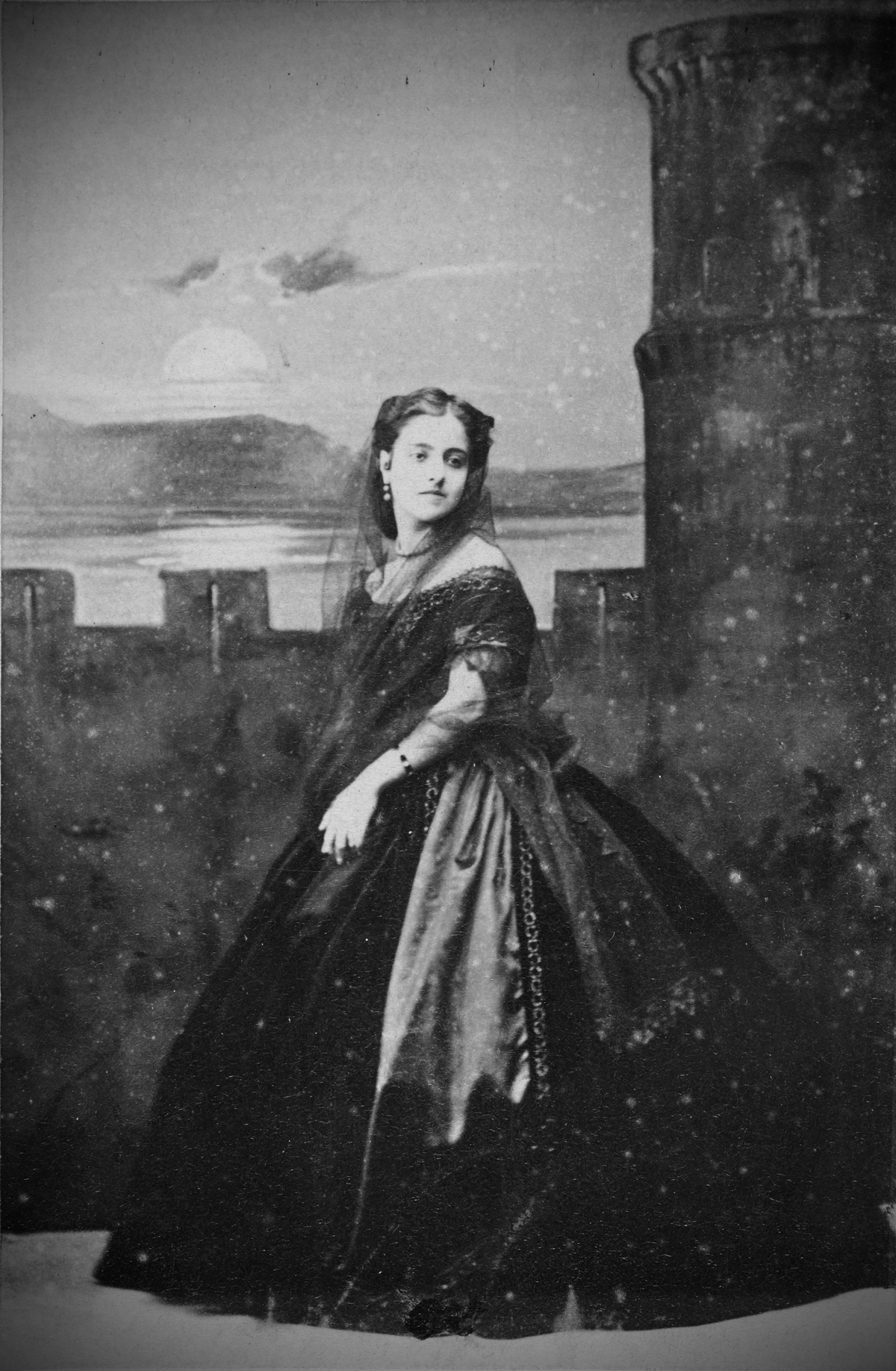 A woman in a dark, elegant 19th-century dress stands outdoors near a stone tower, with a sunset and water in the background. She has dark hair and wears jewelry, looking slightly to her left.