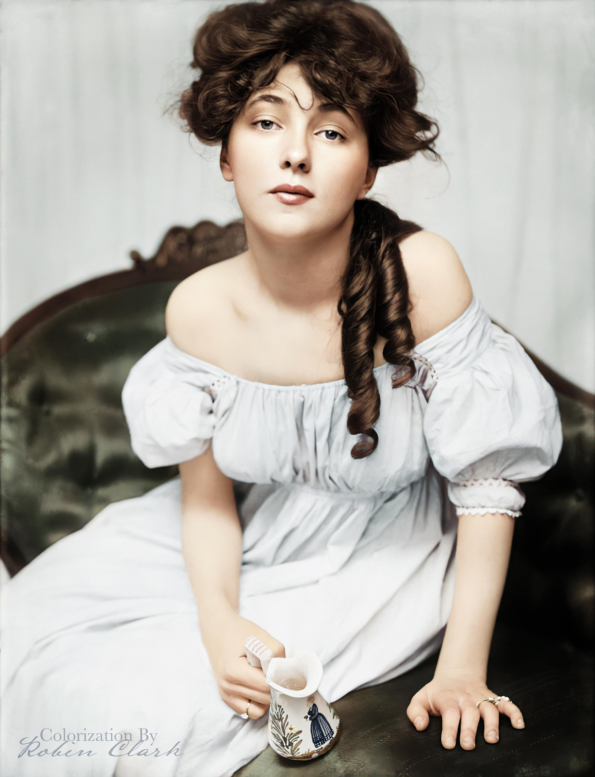 A young woman in a white off-shoulder dress sits on a vintage chair, holding a small decorated jug, with her long, curly hair draped over one shoulder.