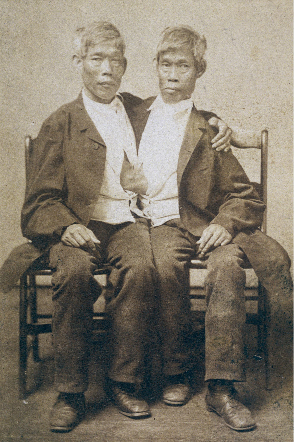 Two men sit side by side on a wooden bench, joined at the torso. They wear similar dark jackets, light shirts, and pants, and one has his arm around the other. The photo is sepia-toned and vintage.