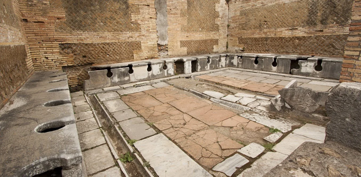 Ancient Roman public latrine with stone benches featuring keyhole-shaped openings, set against brick walls and a stone-tiled floor, showing historic communal restroom design.