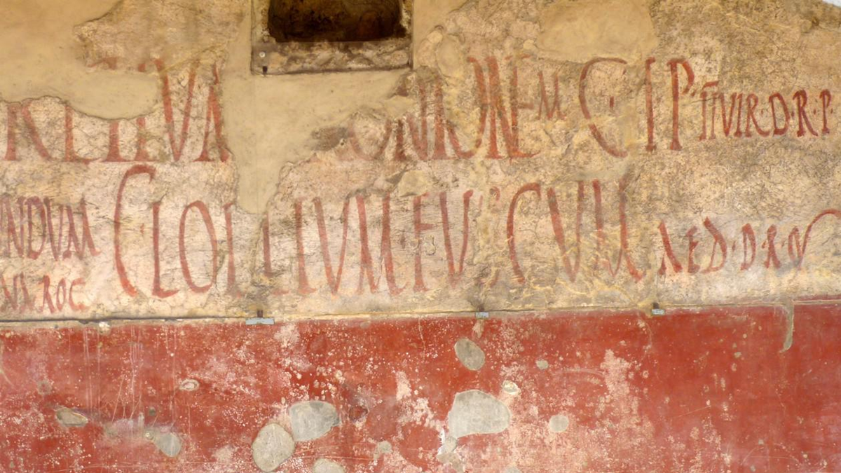 Ancient red Latin graffiti painted on a faded, cracked wall with a red lower section and beige upper section. Some letters are missing or incomplete due to wall damage.