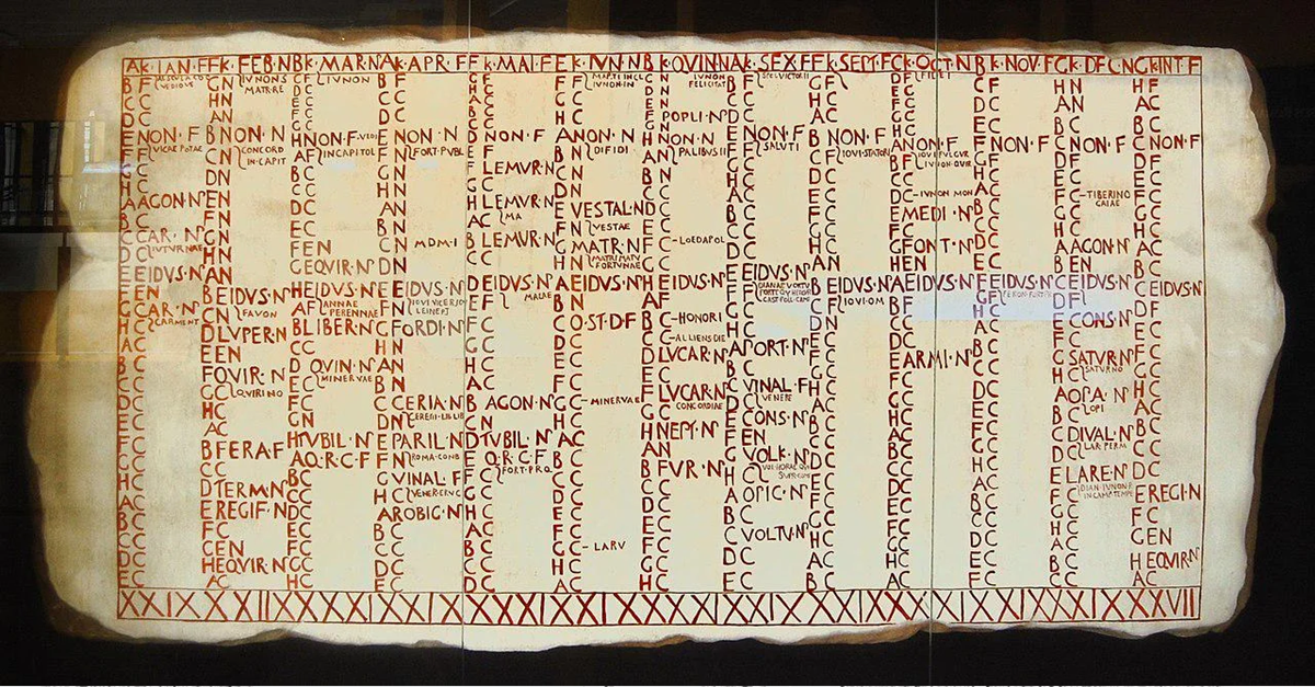 A large parchment features a grid filled with Latin letters and abbreviations in red and black ink, bordered by two rows of red Roman numerals along the top and bottom edges.