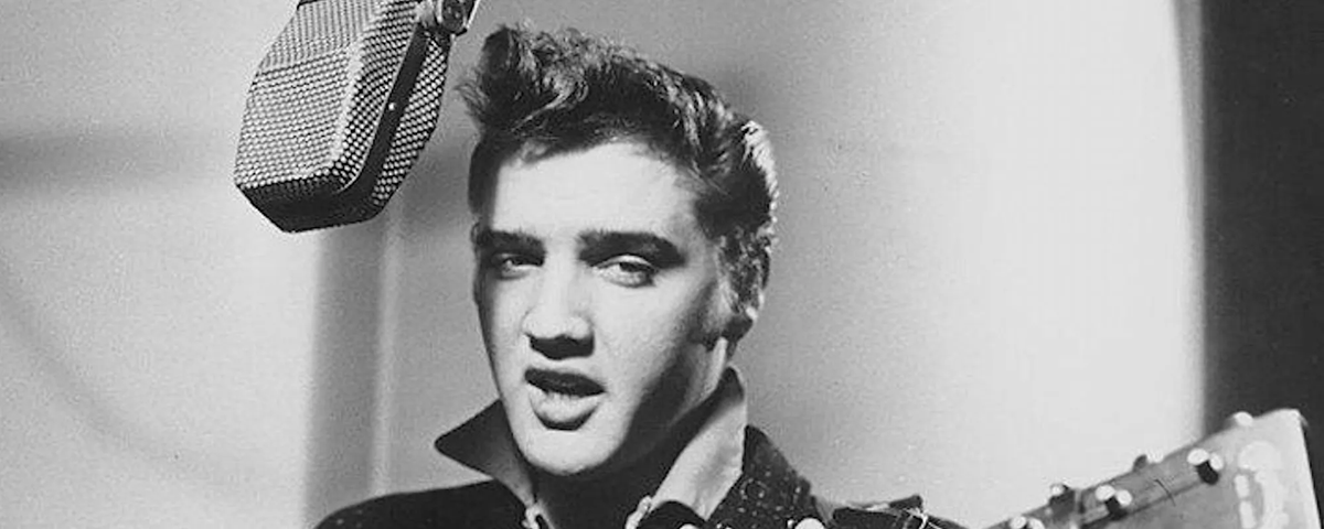 A young man with styled dark hair sings into a large vintage microphone while playing guitar. He wears a collared shirt and looks slightly to the side in this black-and-white photo.