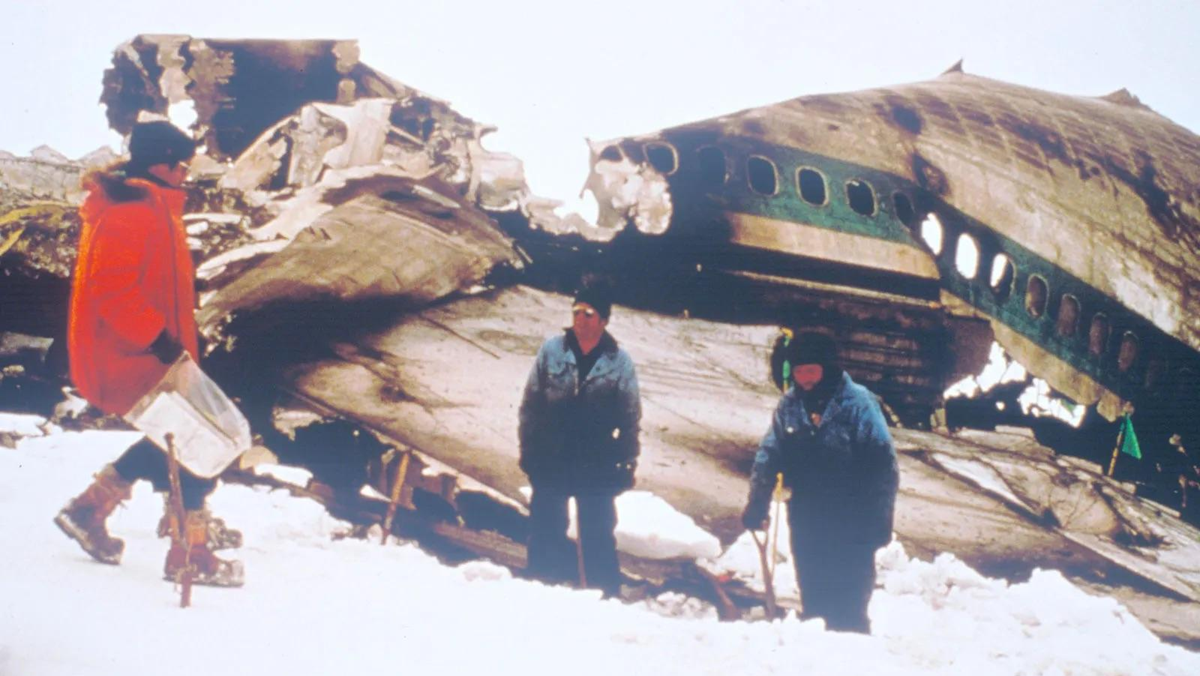 Three people in winter clothing stand in snow near the wreckage of a large airplane. The broken fuselage is visible, with holes and torn metal, indicating a crash in a snowy, cold environment.