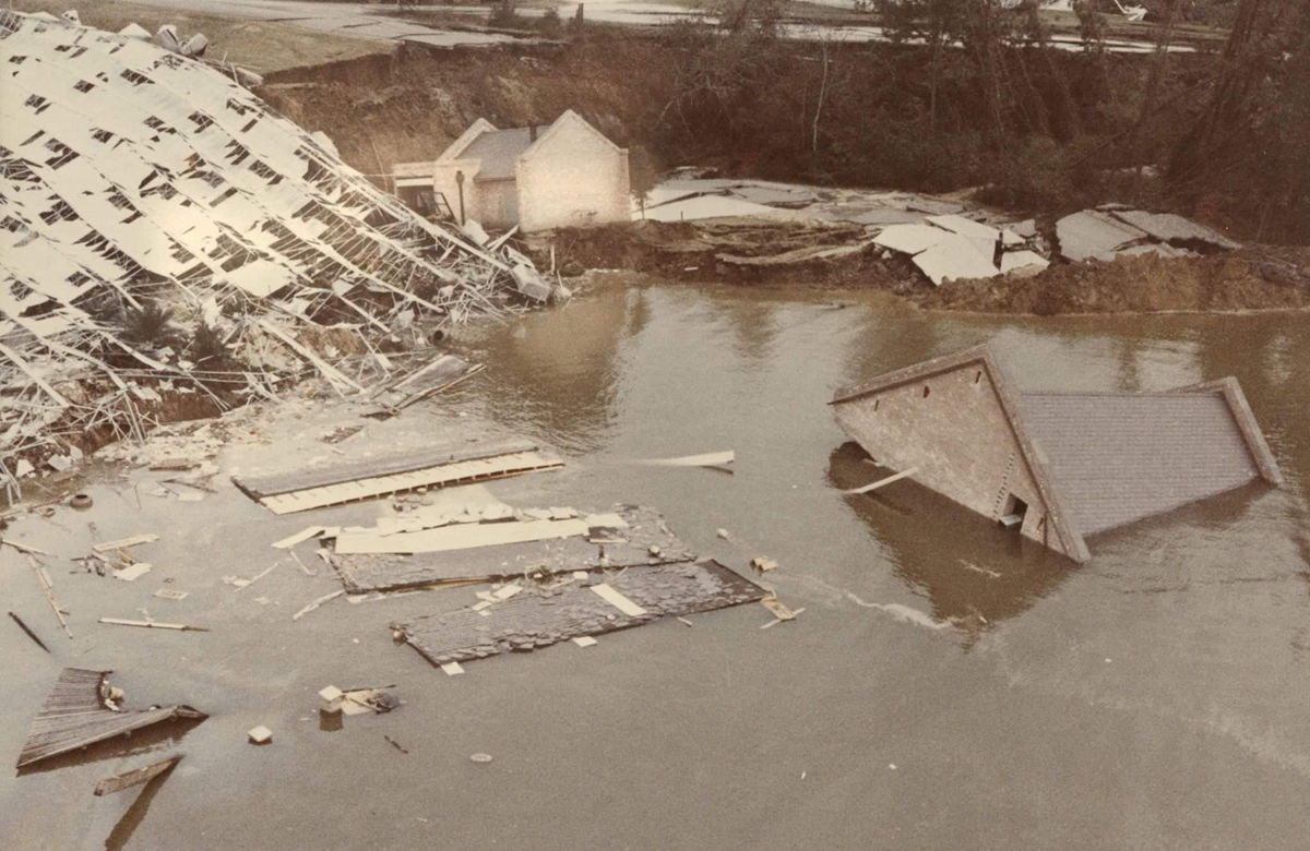 A flooded area with collapsed buildings, debris floating in the water, and partially submerged structures near a muddy shoreline.
