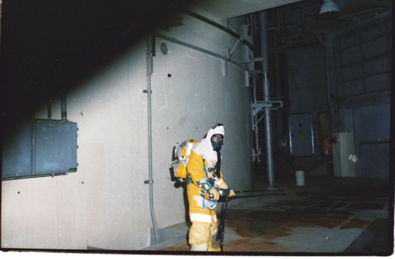 A person in a yellow hazmat suit and respirator stands in an industrial facility with pipes and large doors visible in the background.