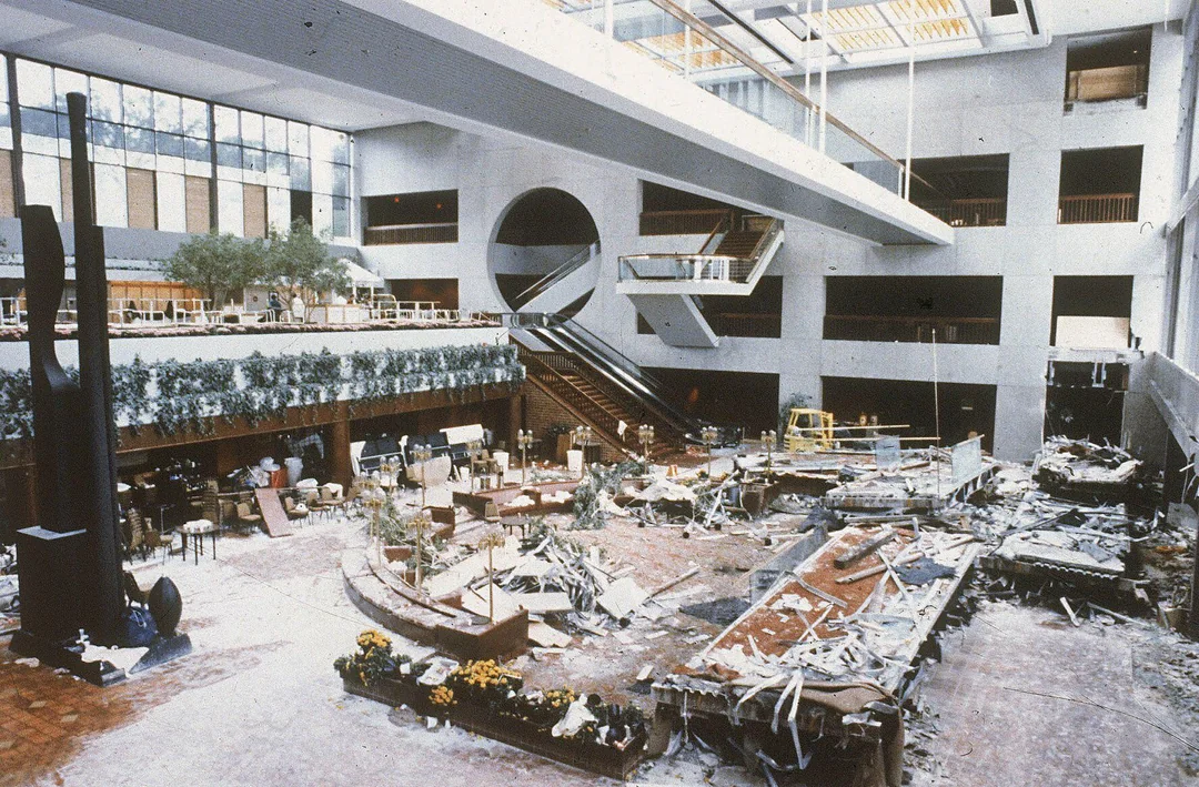 An indoor atrium with visible damage and debris scattered across the floor, broken furniture, and construction materials, while upper levels appear mostly intact.