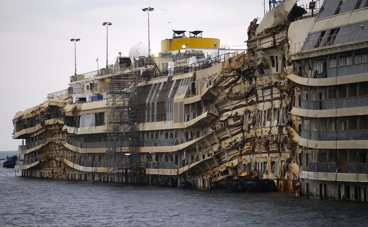 A large cruise ship with a rusted, heavily damaged hull sits partially submerged in water, surrounded by scaffolding for repairs. The upper decks remain mostly intact, contrasting with the twisted lower structure.