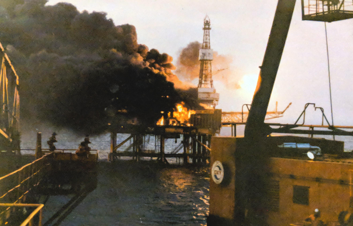 A large fire with thick black smoke engulfs an offshore oil platform at sea, while part of the structure remains visible against the sky and water.