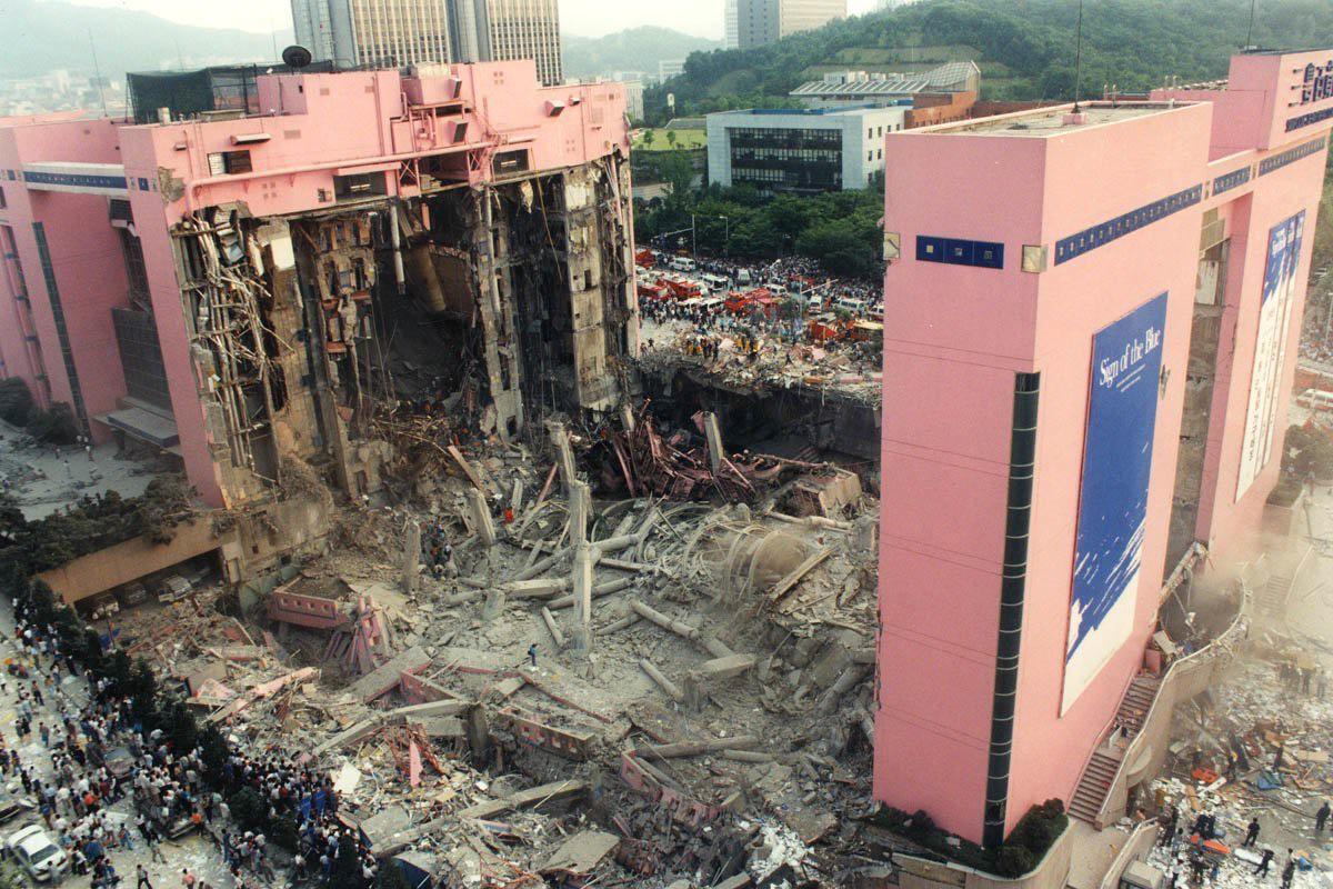 Partially collapsed pink building with large section destroyed, rubble covering the ground, and crowds gathered nearby; other buildings and trees are visible in the background.