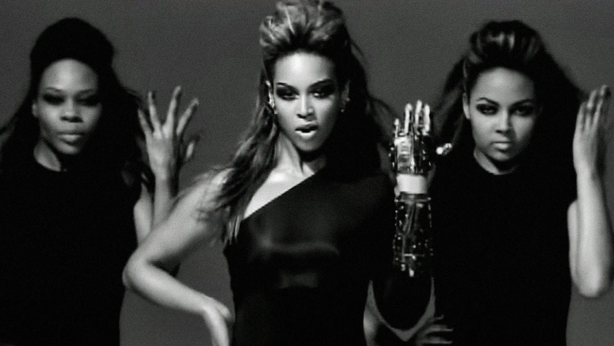 Three women in black outfits perform a dance routine against a plain background. The central woman stands confidently with one arm raised, while the others pose with expressive hand gestures. The image is in black and white.