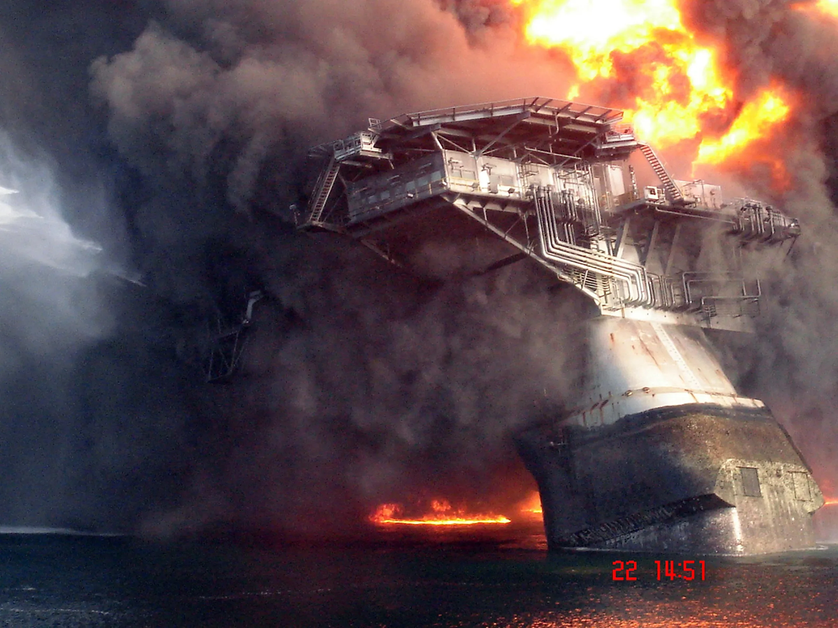 A large offshore oil platform is engulfed in flames and thick black smoke, partially collapsed into the ocean, with fire visible on the water's surface.