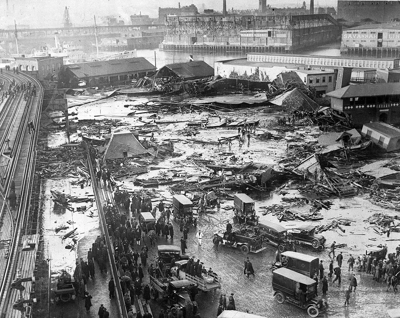 Black and white photo of a city street flooded with debris and people. Old cars and crowds gather near collapsed buildings, with industrial structures and a river in the background.