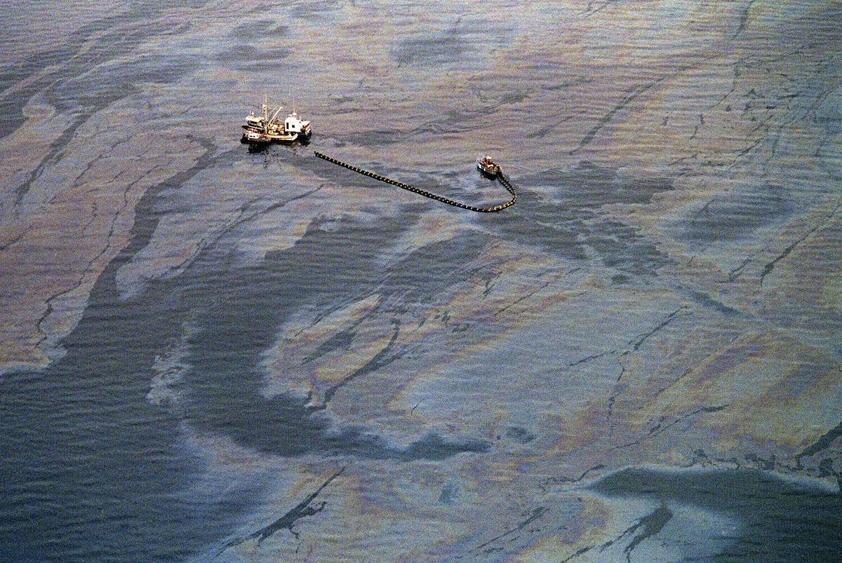 Aerial view of two boats in an oil-polluted ocean, with one towing equipment through dark, slick water.