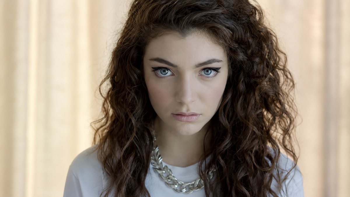 A young woman with long, curly brown hair and striking blue eyes stares directly at the camera. She wears a white top, a chunky silver chain necklace, and has a neutral expression. The background is softly blurred.