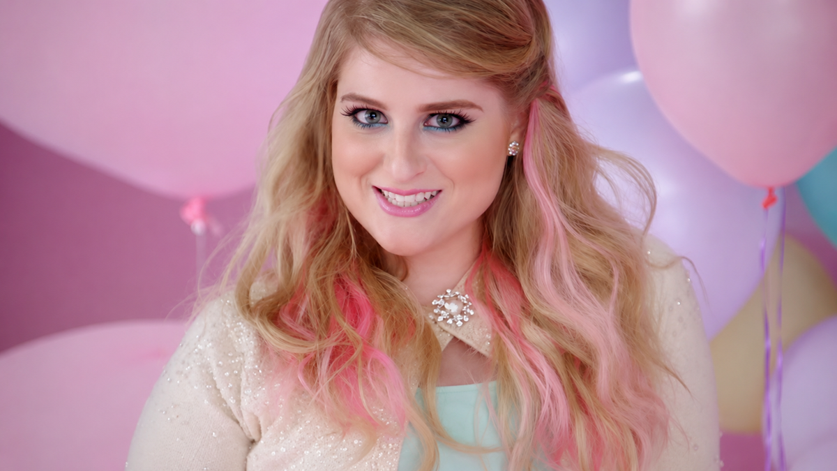 A woman with long blonde hair and pink highlights smiles in front of pastel-colored balloons. She wears a sparkly cream top, a jeweled necklace, and light makeup, with a soft pink and purple background.