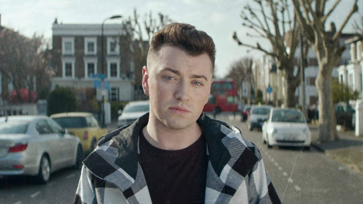 A person with short brown hair wearing a black and white plaid jacket stands on a city street lined with cars and trees on a bright day, with houses and a red double-decker bus in the background.