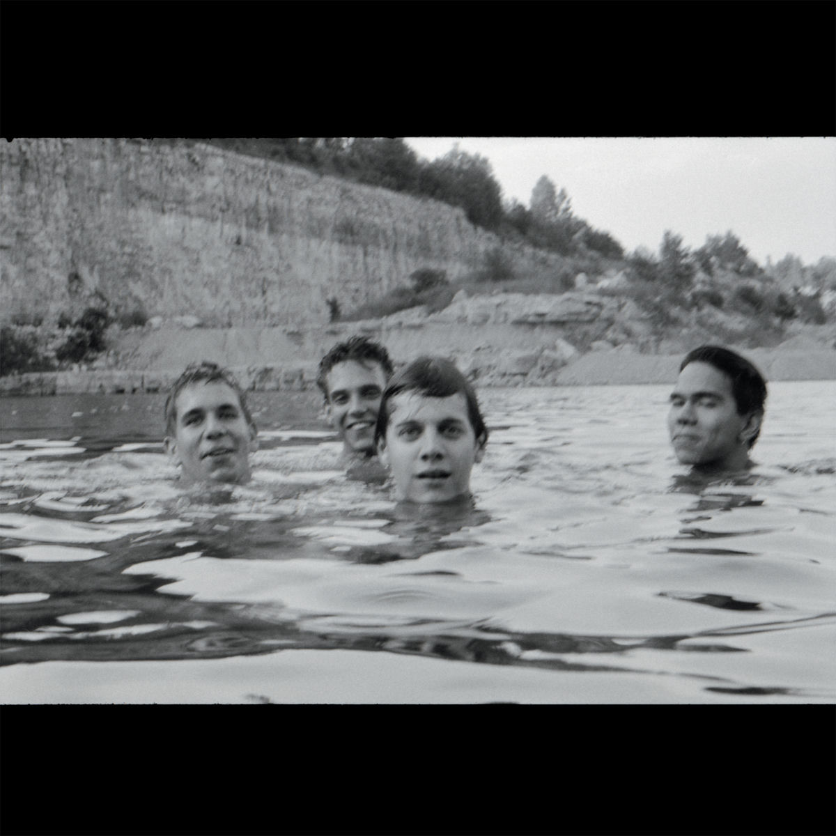 Four people are partially submerged in water, facing the camera. Behind them is a rocky shoreline with trees. The image is black and white and framed with black borders at the top and bottom.