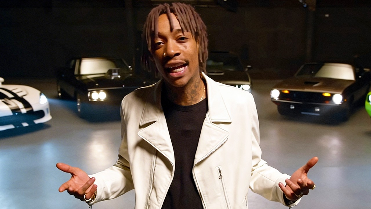 A person with dreadlocks wearing a white jacket stands indoors, gesturing with both hands. Several classic and modern cars are parked in the background under dim lighting.