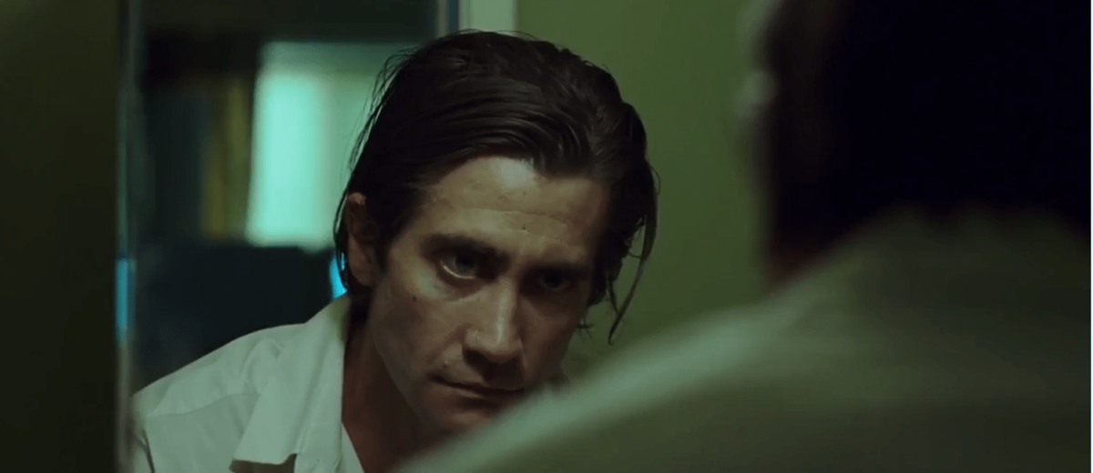A man with slicked-back hair and a serious expression stares intently at his reflection in a mirror, set against a dimly lit, green-toned background.