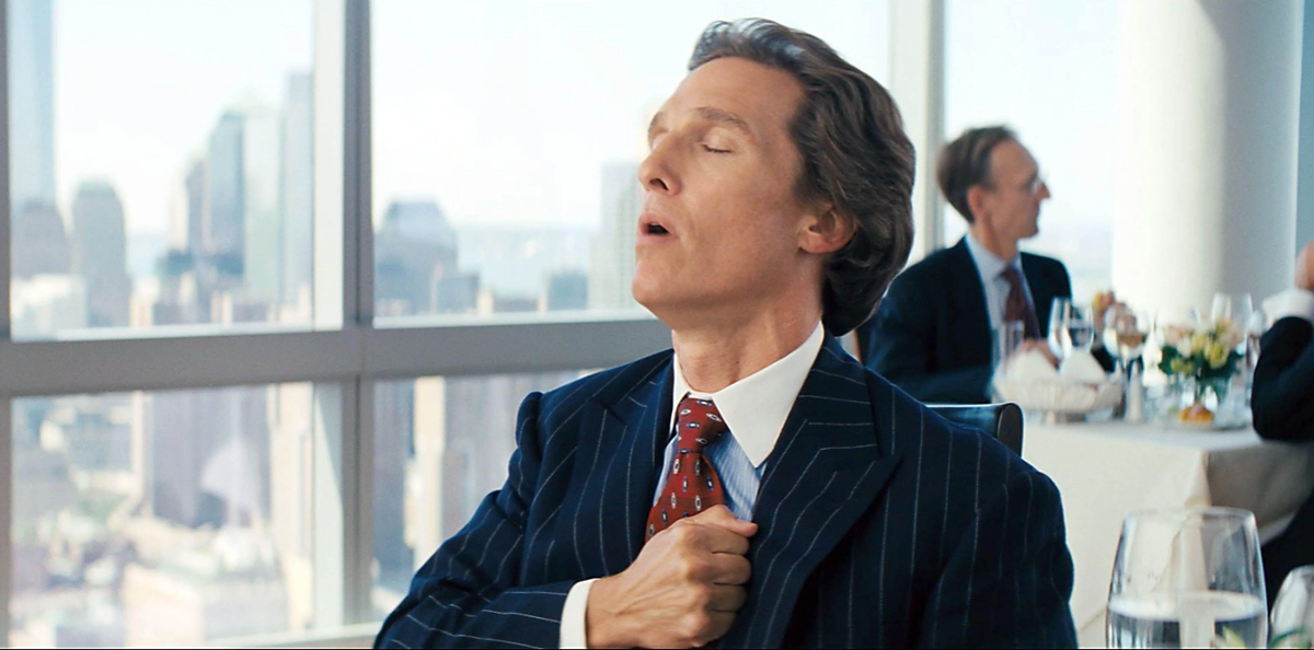 A man in a pinstripe suit sits at a restaurant table, eyes closed, and thumps his chest with his fist. City buildings are visible through large windows in the background.