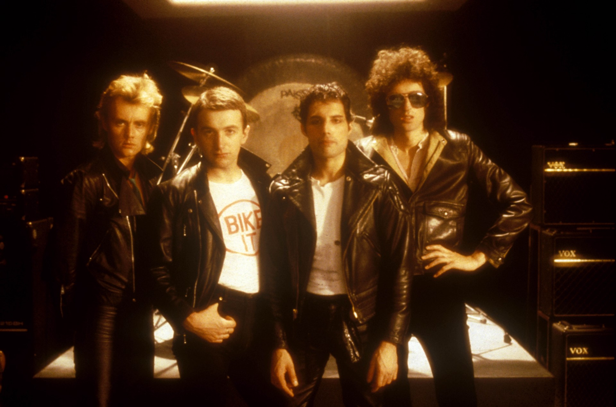 Four men stand in front of drum sets and amplifiers, wearing black leather jackets and posing confidently in a dimly lit room, resembling a classic rock band publicity photo.
