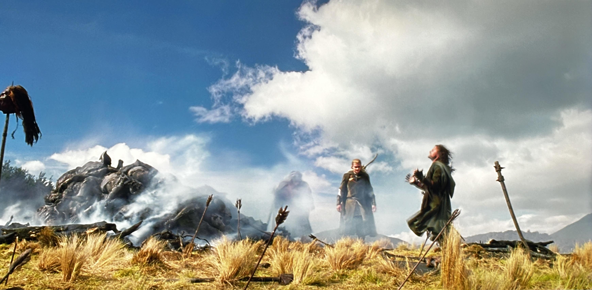 Two people stand in a smoky field with tall grass, surrounded by spears. A fallen creature lies in the background under a blue sky with scattered clouds.
