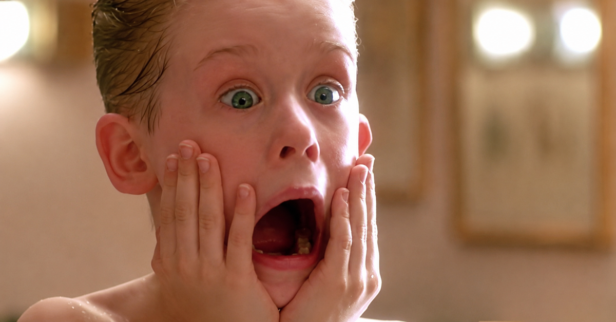 A young child with wet, slicked-back hair holds both hands to his cheeks and looks shocked with wide eyes and mouth open, in a bathroom setting.