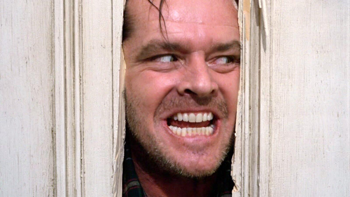 A man with a wild expression peers through a hole in a broken wooden door, grinning intensely with teeth bared.