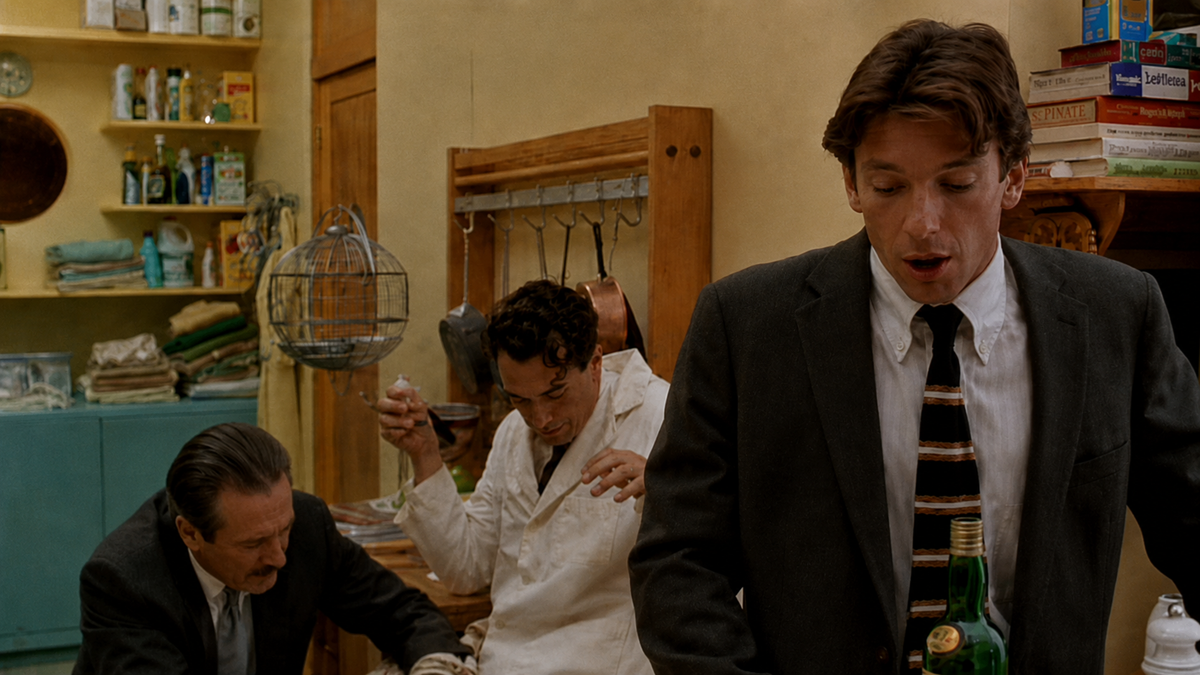 Three men are in a cluttered kitchen; one stands in a suit pouring a drink, while two others sit at a table, one in a white coat eating and the other looking down. Shelves with books and kitchen supplies are in the background.