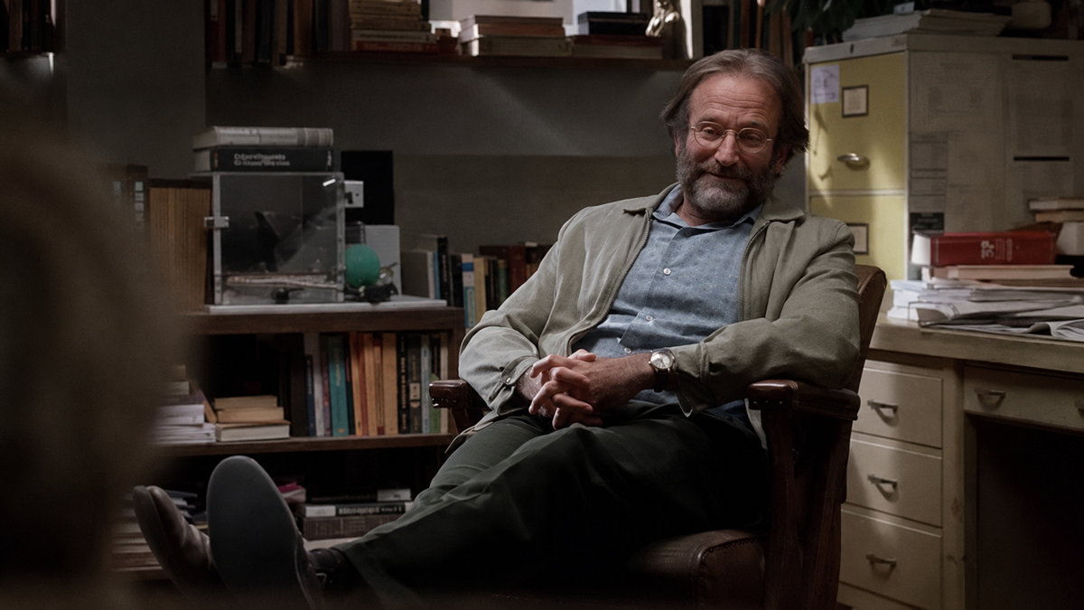 A man with glasses and a beard sits relaxed in an office chair, hands clasped, surrounded by books, papers, and a filing cabinet, appearing to listen attentively.