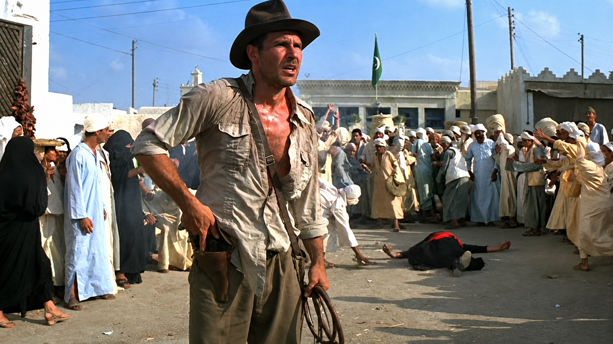 A rugged man in a fedora and dusty clothes stands alert in a crowded marketplace as people in robes look on, some in shock. A person lies on the ground behind him.