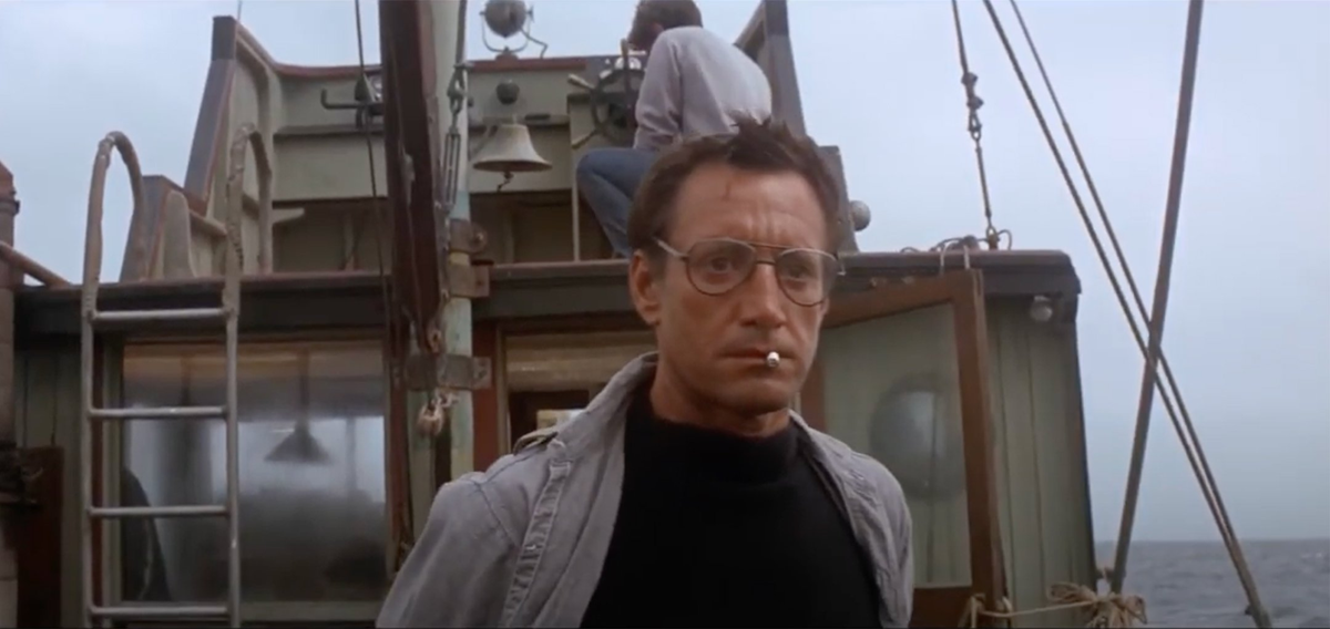 A man wearing glasses and a cigarette in his mouth stands on a boat deck, looking serious. Another person is seated behind him at the boat's controls. The ocean is visible in the background.