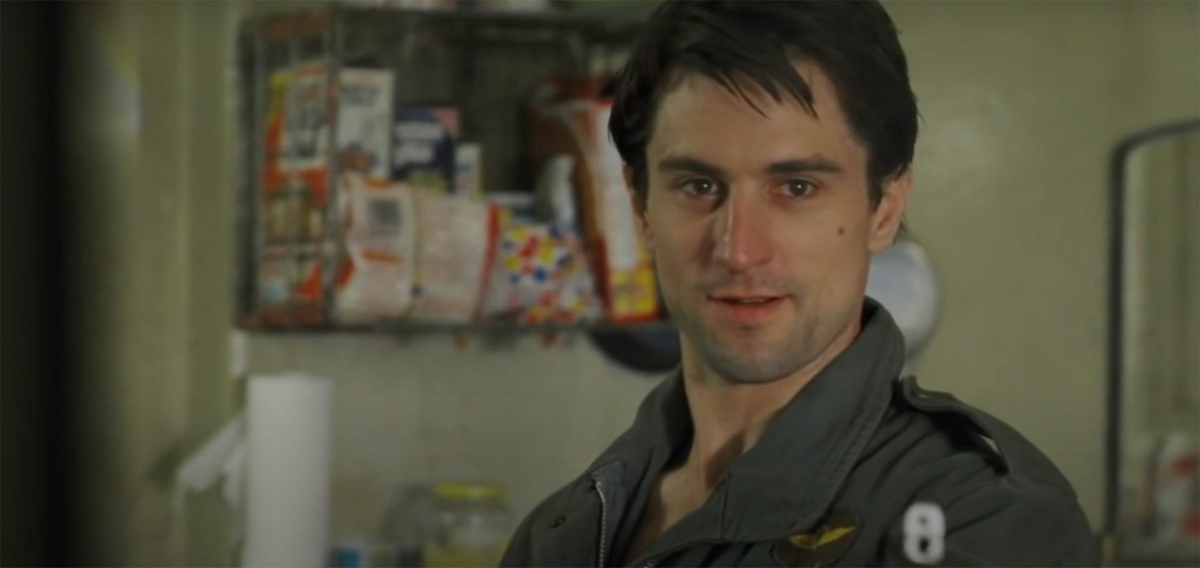 A young man with dark hair in a green jacket stands in a kitchen, looking forward with a slight smile. Shelves with food items and a paper towel roll are visible in the background.