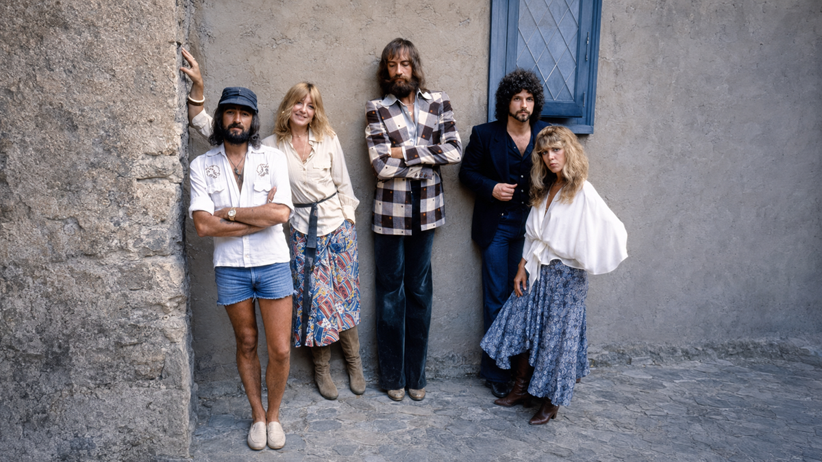 Five people stand against a stone wall outside, dressed in 1970s-style clothing, including skirts, boots, shorts, and patterned shirts, with relaxed and casual poses.